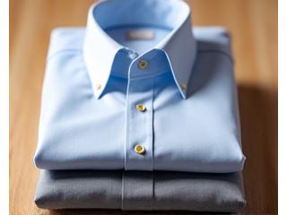 Tumpukan kemeja pria rapi dalam warna putih, biru muda, dan abu-abu di atas meja kayu dengan cahaya alami.